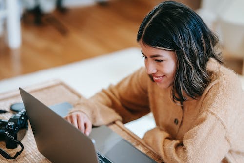 Free Smiling Latin American female remote employee working on netbook at table with photo camera in house Stock Photo